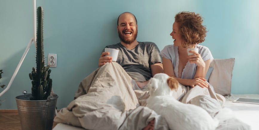 Una pareja blanca pelirroja pasa la mañana en la cama, riendo y tomando café contra una pared azul con un perro blanco abrazado en el edredón.