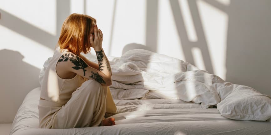 Side view of a white woman with red hair sitting on a bed covered in white linens against a white wall splattered with sunlight, covering her face with her hands.