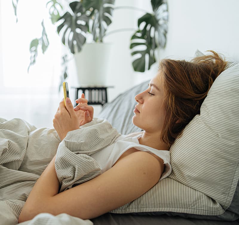 Young woman holding a yellow phone, in bed, tapping on her phone