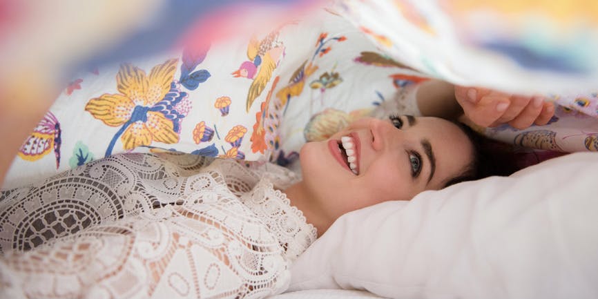 Una joven blanca en la cama se esconde debajo de una sábana, sonriendo mientras mira la sábana del color del arcoíris sobre ella.