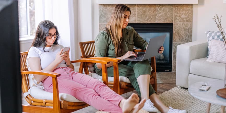  A young Latina teen sits in a chair looking into her phone, with a female relative sitting in a chair next to her, working on a laptop.