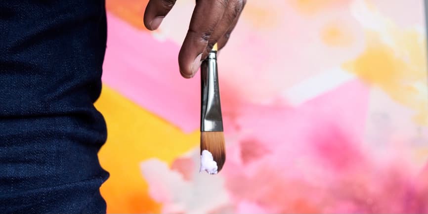 A close-up shot of a Black man’s hand, holding a paintbrush dipped in white paint, against a pink pastel backdrop.