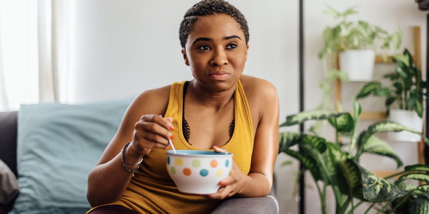 Una joven negra con el pelo corto y una camiseta amarilla sin mangas se sienta en un sofá, comiendo un tazón de cereal en un tazón colorido.