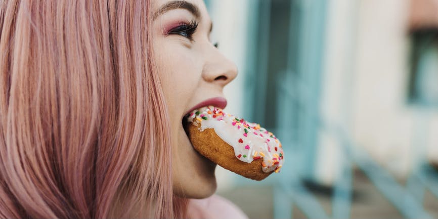 A beautiful young white Asian woman with pink hair holds a donut topped with sprinkles between her teeth while smiling and looking away from the camera. 