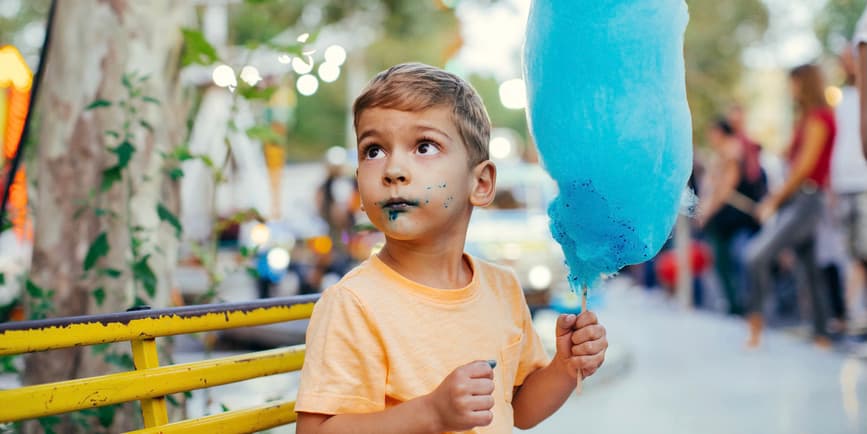 A young white toddler sits on an amusement park bench holding a giant bright blue cotton candy. He’s looking off camera with a blue stain on his mouth.