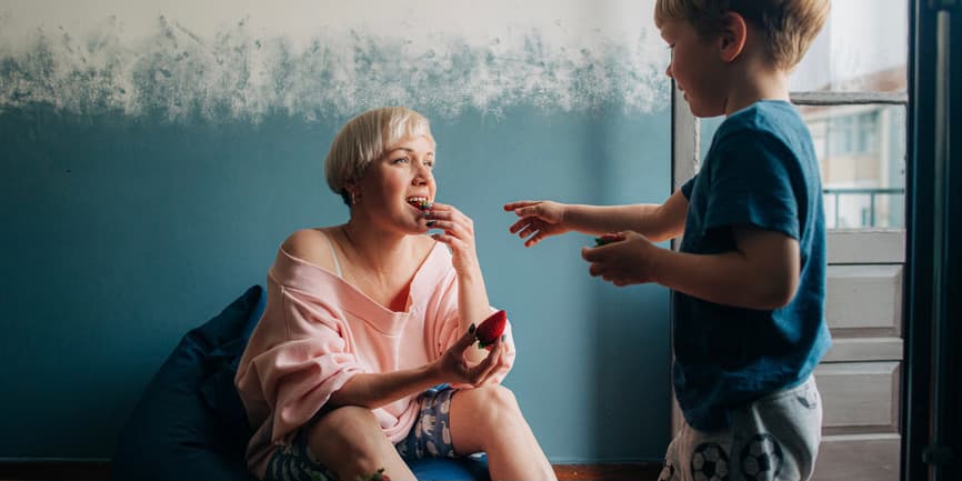 A young white mother with short blond hair wearing a pink sweatshirt sits on a beanbag against a blue wall. She’s sharing strawberries with her 5-year old son. They have short blond hair and wear a blue t-shirt.