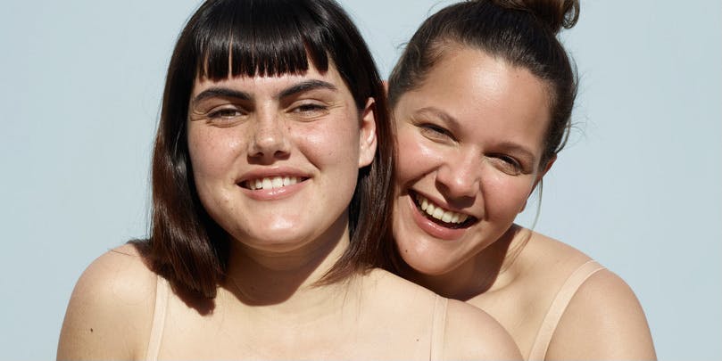 Dos jóvenes amigos blancos posan en ropa interior mientras se divierten bajo el sol. Tienen el pelo oscuro, sonríen, no usan maquillaje y usan colores neutros.