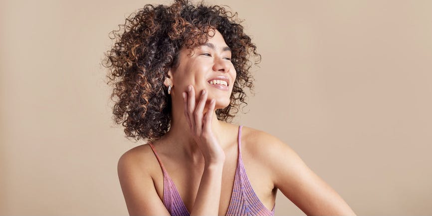 Una joven de piel morena y cabello afro castaño sonríe mientras se toca la mejilla y mira fuera de cámara. Lleva una camiseta sin mangas morada y está de pie contra una pared de color neutro.