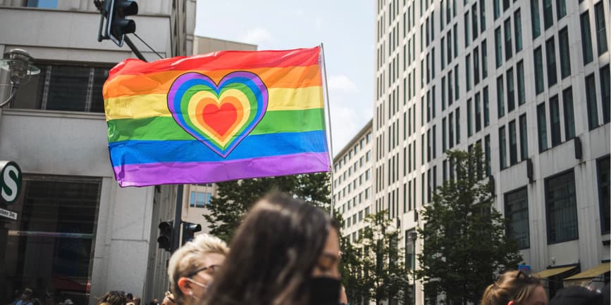 An outdoor photograph of city buildings and the heads of people marching in a Pride parade, with a pride flag with a heart at the center above the crowd.