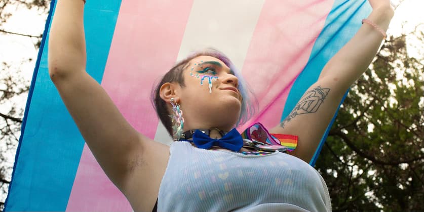 A color photograph of a trans person wearing a white tank top over a colorful body suit holds a Trans Pride flag over their head, smiling gently as they look away from the camera.