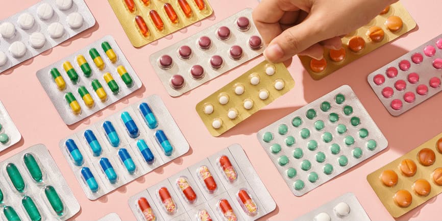 Different types of pills, Tablets in packs, and blister packs with colorful medicines are set on a pink background, with the fingers of a white hand picking one up.