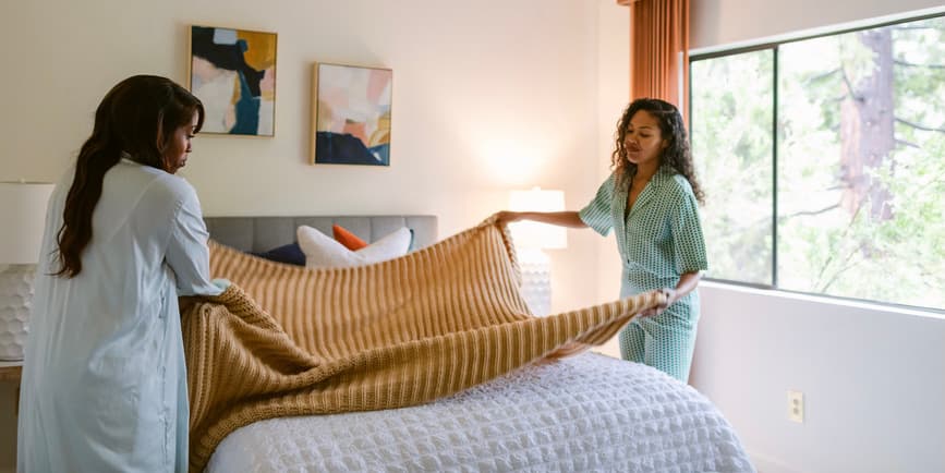 Two Black women wearing pajamas make a bed in a sun-filled room.