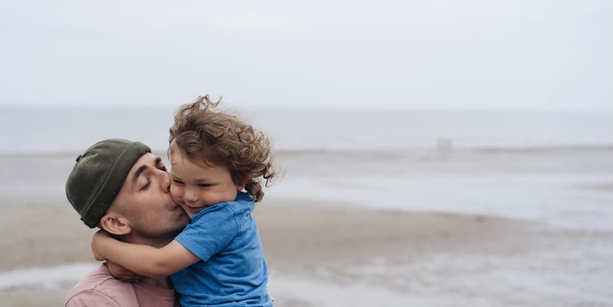 A young white father with shorn hair wearing a warm cap holds his child up close and hugs them.