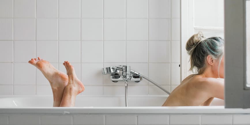 An older white woman with blond and blue hair tied up lays in a bathtub in a white bathroom looking out the window.