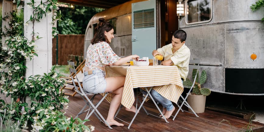 A young white couple sits outside a steel camber van, having a meal on a cloth-covered folding table.