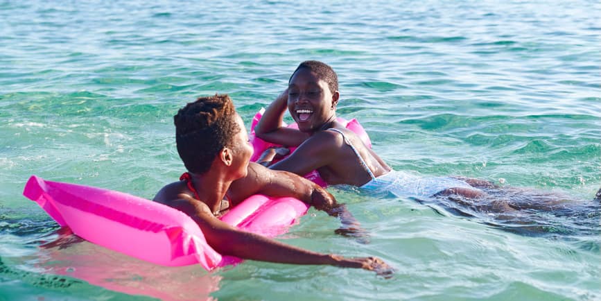 Two laughing young African female friends holding on to a float while swimming together in the ocean on a sunny day.