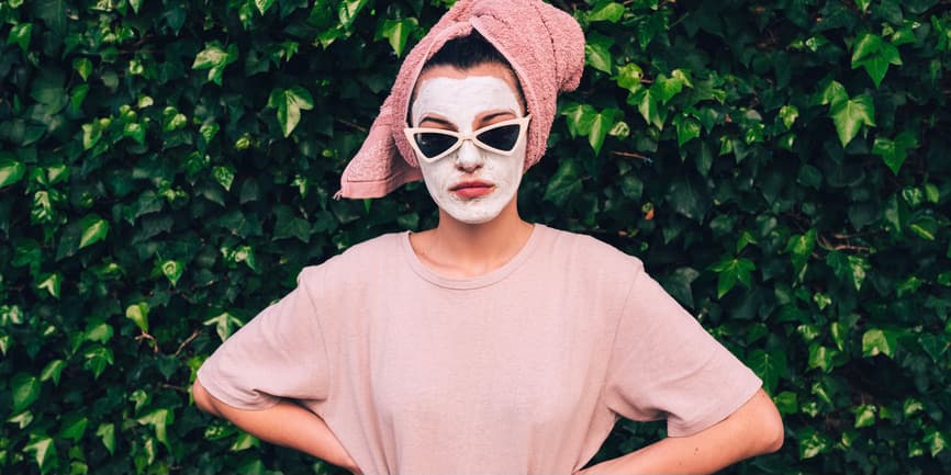 A young white woman with wearing a white skincare face mask, sunglasses, and a towel on her hair, stands against a wall of ivy and looks at the camera