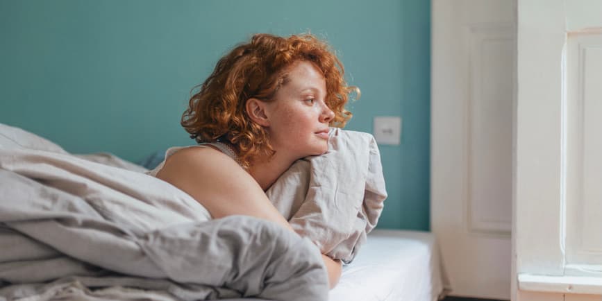 Portrait of a beautiful young white woman with red hair laying on her bed, looking out the window 