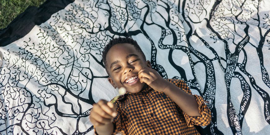 Color overhead photo of a Black toddler wearing a brown shirt lying on a blanket in the grass laughing and looking at the camera while holding a small flower in his hand