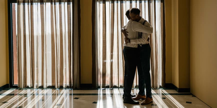 A sun-dappled photo of two young, thin Black men wearing dress pants and white shirts, hugging in front of a large draped window.