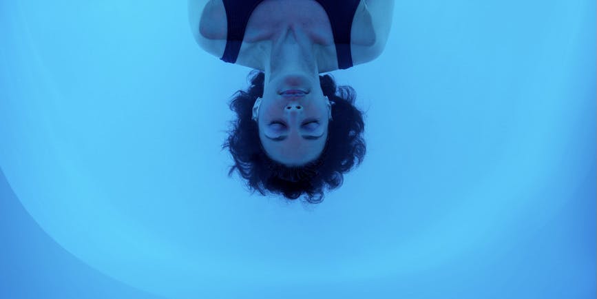 A young white woman floats with her eyes closed in the blue water of a sensory deprivation tank.