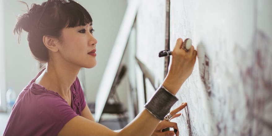 An Asian woman wearing a purple shirt with her brown hair tied up is concentrating on drawing on her canvas with a pen. The artist is working in a studio with daylight.