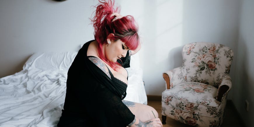 Una mujer curvilínea con cabello rosado, lencería y un suéter negro se sienta en su cama con la cara inclinada hacia abajo.
