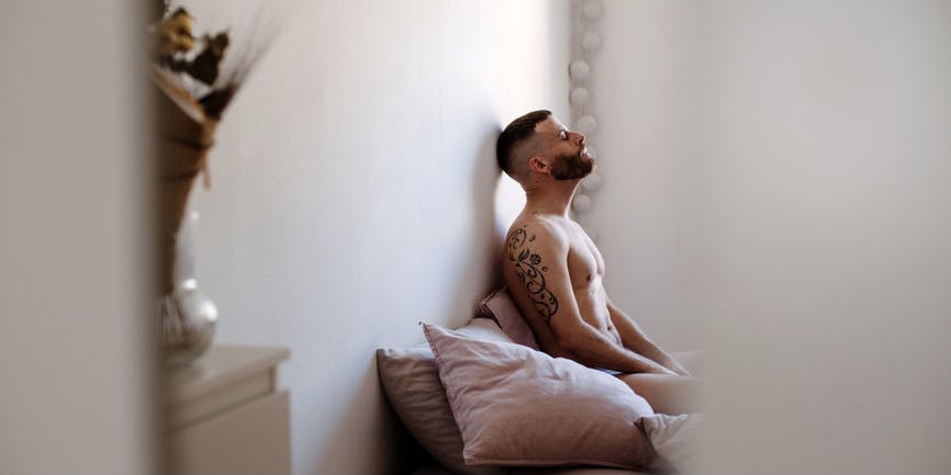 A white man with short hair and a beard wearing no shirt sits in bed with his head up against the wall. He has tattoos on his arms and is viewed through the doorframe. 