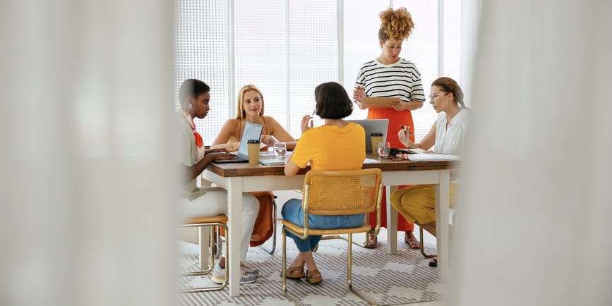 Grupo de compañeras vestidas de manera casual sentadas a la mesa con un portátil y discutiendo los detalles del proyecto mientras trabajan juntas en un espacio de trabajo conjunto