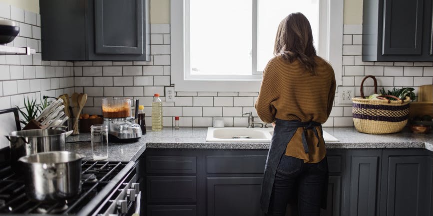 A white woman with blond hair wearing a brown sweater is viewed from behind washing dishes at a sink in a bright kitchen with black cabinets. 