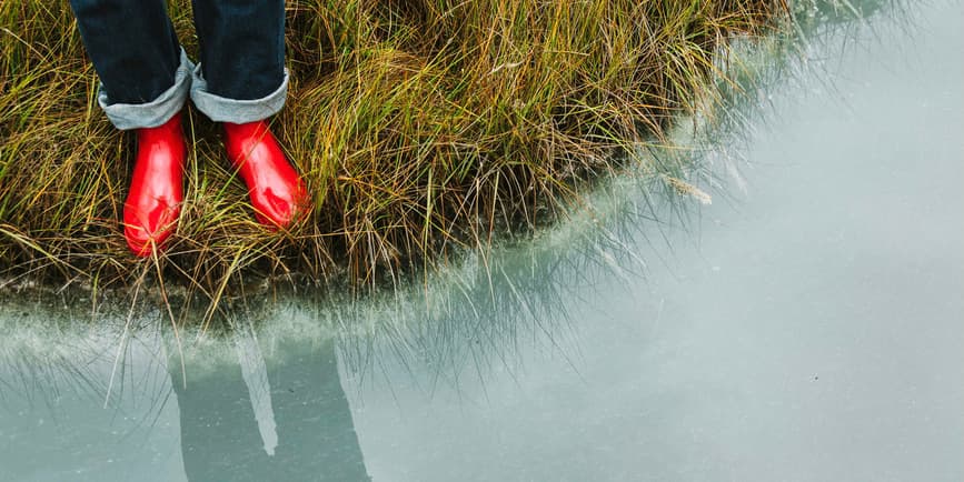 An overhead shot of the legs of a person wearing dark blue jeans and red rubber boots standing in the grass at the edge of still misty-blue water.