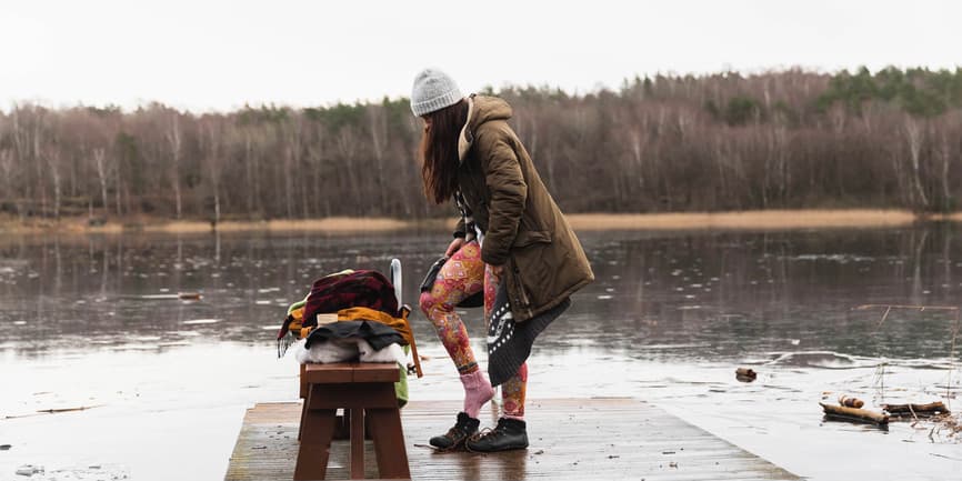 A white woman with long brown hair wearing a winter coat, hat, leggings and boots stands on a dock in the middle of winter, preparing to get into the water.