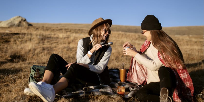 Two young girls white girls wearing hats and warm clothes picnic outside in a brown field during fall.