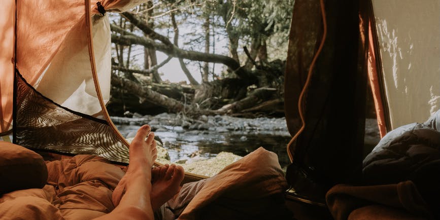 A color photograph from the inside of a camping tent, with the flap open showing a woodland picture outside and the legs of someone layout out on a sleeping bag within.