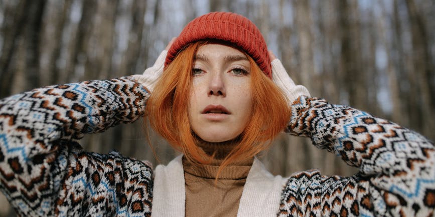 Portrait of a white woman with red hair wearing a sweater and red hat, looking into the camera as they stand outside in front of bare trees.