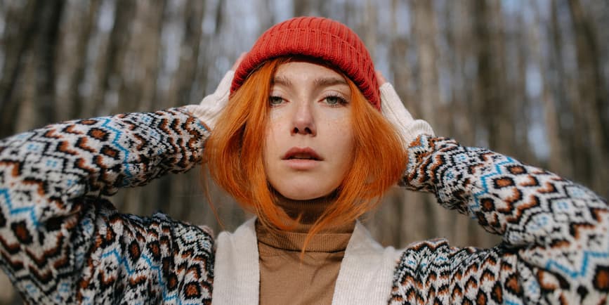 Portrait of a white woman with red hair wearing a sweater and red hat, looking into the camera as they stand outside in front of bare trees.