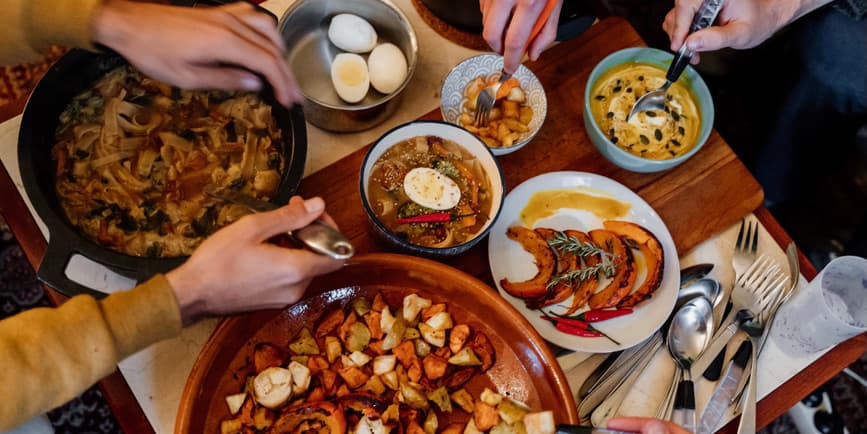 Overhead photograph of a low table with plates and dishes of autumn foods, and several hands putting utensils into the dishes.