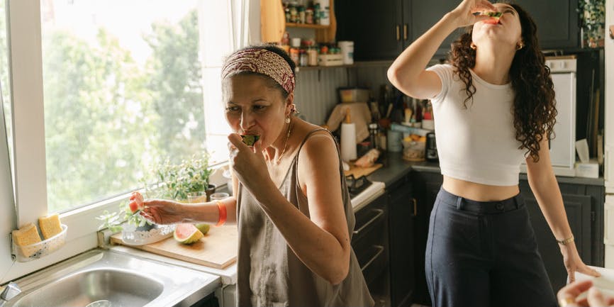 A Latina mother and adult daughter eating watermelon in their kitchen. The kitchen is full and cluttered, with lots of beautiful colors/