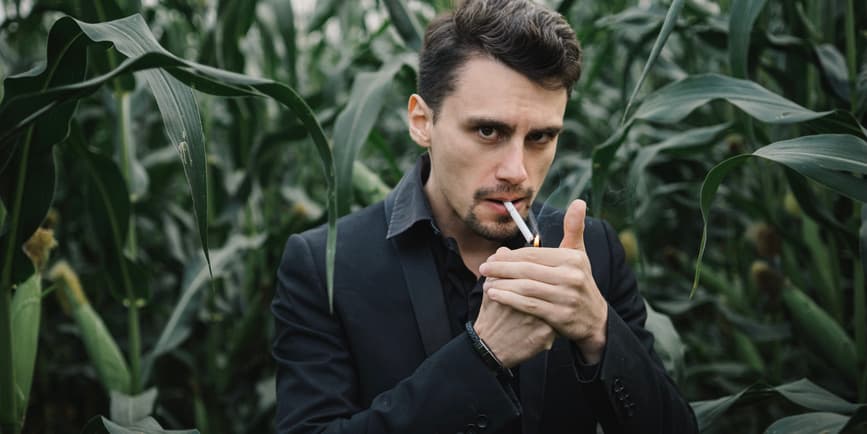 A young white man with short brown hair dressed in black suit stands in a corn field and looks at the camera as he lights a cigarette.