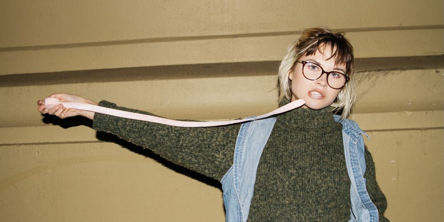 A young white teenager with short blond hair wearing black glasses, a green sweater and denim vest has the end of a roll of gum tape in her mouth and pulls the rest away from her.