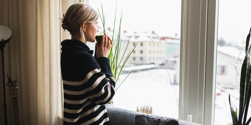 A white woman with long blond hair tied back wearing a striped sweater stands looking out a window while sipping from a glass.