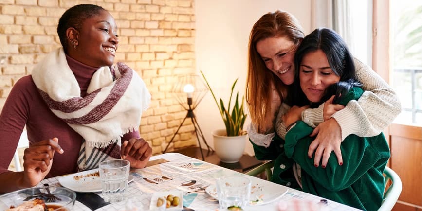 Female friends in winter clothing gather at a table, smiling and hugging each other.