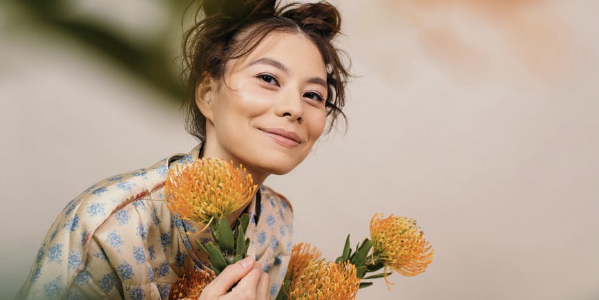 A cheerful young Asian woman with her brown hair tied up wears a stylish colorful robe and holds orange flowers as she smiles gently into the camera. 