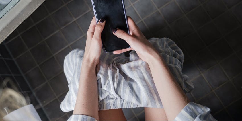 Foto desde arriba de las piernas y manos de una mujer sujetando y enviando mensajes de texto a su teléfono en un baño de azulejos oscuros. Mujer joven lleva un pijama a rayas azules.
