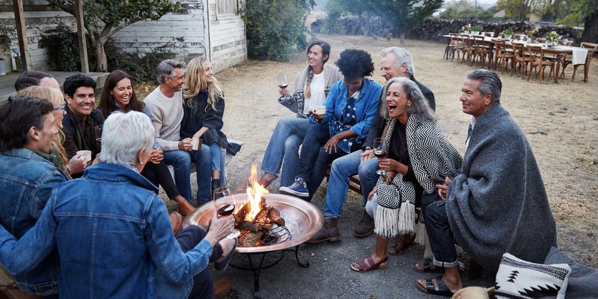 Grupo de familiares y amigos multigeneracionales que se relajan alrededor de una fogata en la región vinícola de Sonoma, en California, después de una cena. Están bebiendo vino y riendo.