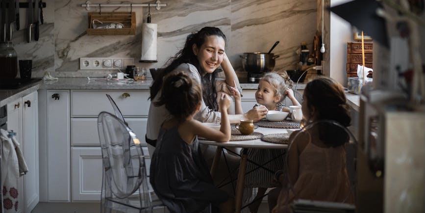 Una madre latina y sus tres hijas pequeñas se sientan a la mesa de la cocina cenando y conversando.