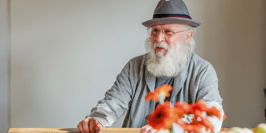 Enchanting grey-bearded white senior man with an elegant style looks away from the camera and smiles while sitting at a kitchen table at home.