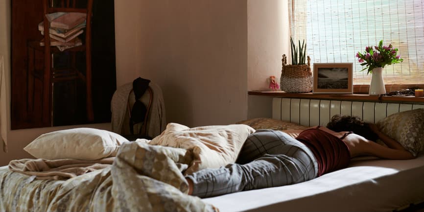 A woman wearing pajamas lies on a bed covered in rumpled blankets, her face down in her pillow, as sunlight streams in through curtains.