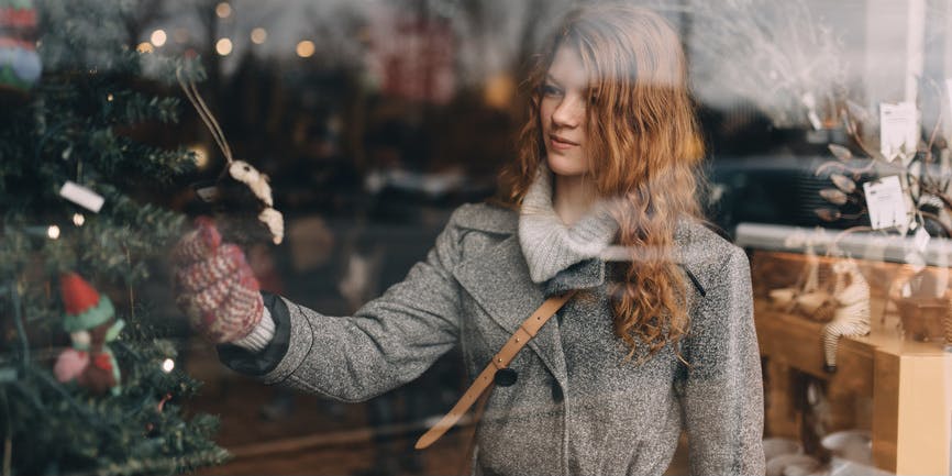 A young woman with long red hair wearing a gray winter coat is seen inside through a shop window handling ornaments dangling from a Christmas tree.
