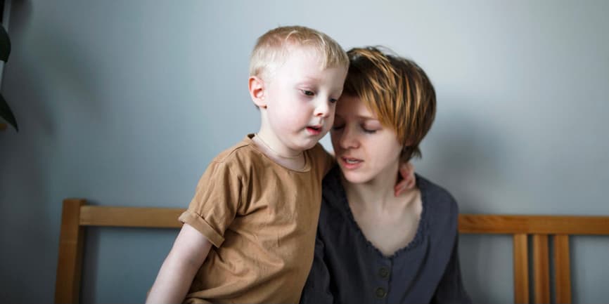 A young white mother with short red hair sits in bed looking tired as she talks to her young blonde son who is gently hugging her.